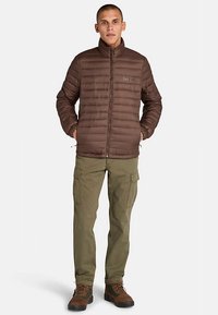 Bruine geïsoleerde pufferjas met horizontale doorgestikte naden, ritssluiting en een klein logo op de borst; gecombineerd met cargobroek en laarzen.