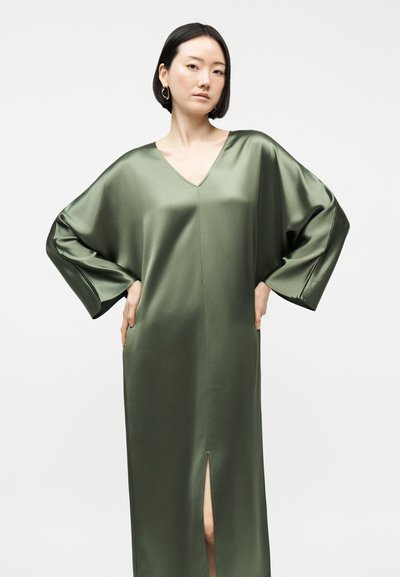 Γυναίκα με κοντά μαύρα μαλλιά φοράει ανοιχτό ξανθό, γυαλιστερό σατέν φόρεμα ελιάς με V-λαιμόκοψη, φαρδιά μανίκια και εμπρόσθια χαραγή, στέκεται με τα χέρια στη μέση.