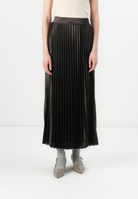 Jupe plissée noire avec une taille haute, fabriquée en matériau brillant. Portée avec des chaussettes grises et des chaussures brillantes à bout pointu.