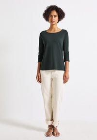Maglietta a maniche lunghe verde scuro, in tessuto morbido, con scollatura rotonda, abbinata a pantaloni beige arrotolati e sandali argentati. Outift semplice e casual.
