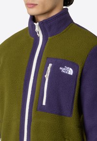 Felpa in pile di colore verde oliva e viola. Presenta un colletto alto, zip completa e una tasca sul petto con zip. Logo bianco sulla tasca.