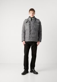 Giacca a vento grigia con tasche grigie a contrasto, chiusura zip a frontale e colletto texturizzato. Abbinata a pantaloni neri e scarpe nere.