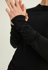 Pull noir en maille avec un col rond et des poignets côtelés, présentant une texture lisse et une coupe ajustée, les mains jointes aux poignets.
