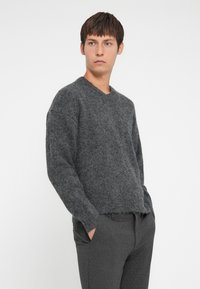Szary, teksturowany sweter z dekoltem w serek i luźnym krojem, zestawiony z szarymi spodniami w kratę. Miękki materiał widoczny w detalach tkaniny.