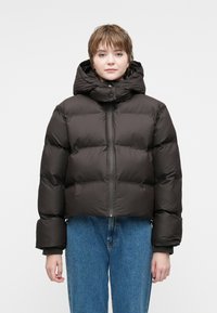 Chaqueta acolchada marrón oscuro con un cuello alto, diseño corto, grandes secciones acolchadas y cremallera en el frente. Combinada con jeans azules.