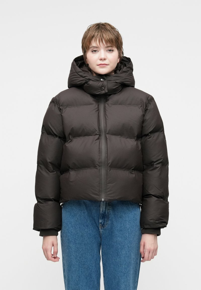 Chaqueta acolchada marrón oscuro con un cuello alto, diseño corto, grandes secciones acolchadas y cremallera en el frente. Combinada con jeans azules.