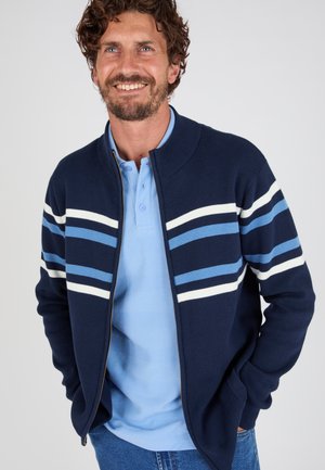 Glimlachende man die een marineblauwe trui met rits draagt, met witte en lichtblauwe strepen over een lichtblauw poloshirt en een blauwe spijkerbroek, staand met zijn handen in zijn zakken.