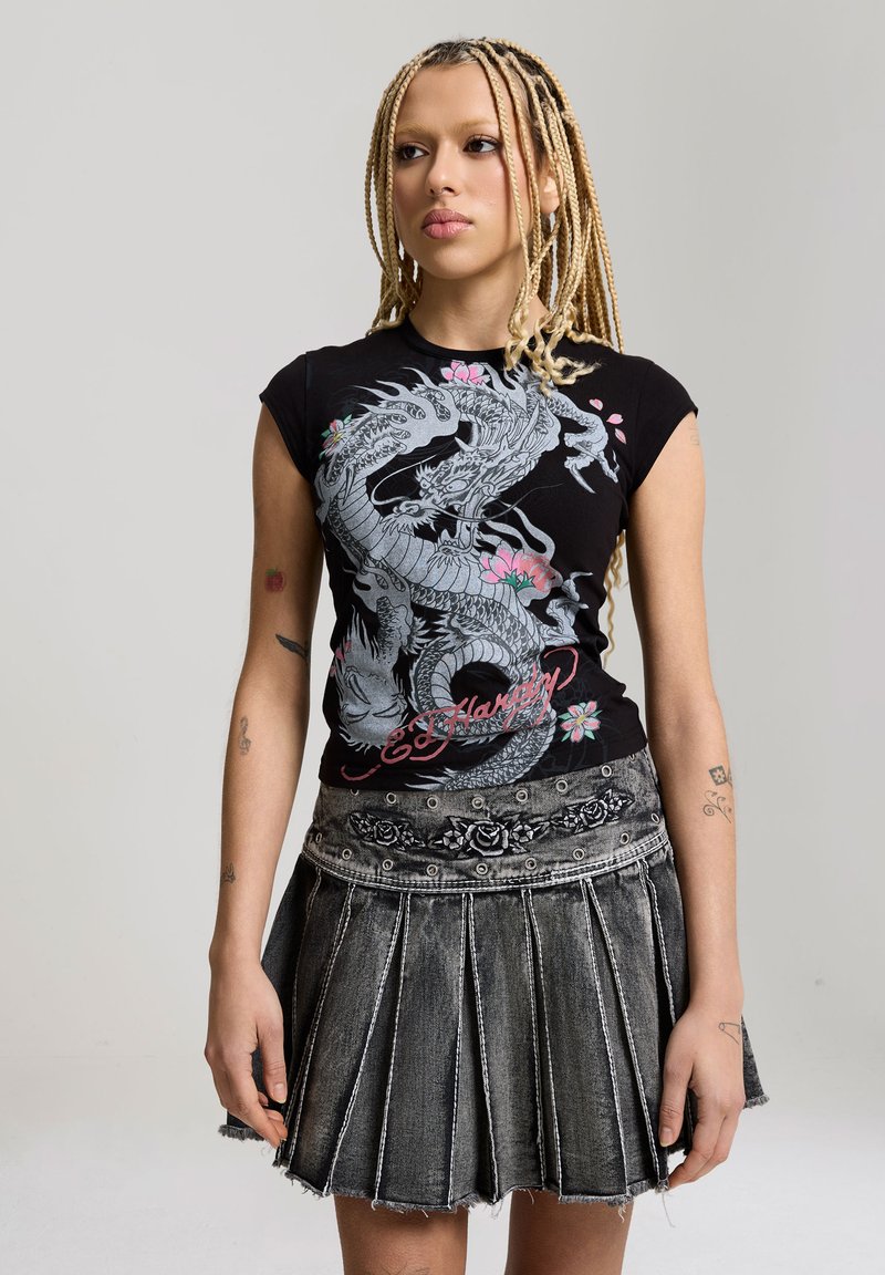Jeune personne aux longues tresses blondes portant un t-shirt noir à motif de dragon et une jupe en jean grise plissée avec une broderie florale.