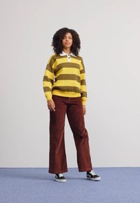 Geel en bruin gestreepte trui met een witte kraag, gecombineerd met bruine corduroy wijde broek en zwarte sneakers op een blauwe vloer.