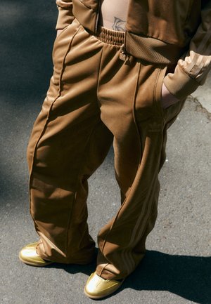 Pantaloni tuta in velluto marrone con vestibilità ampia, con strisce laterali e vita elasticizzata. Abbinati a calzature dorate lucide.