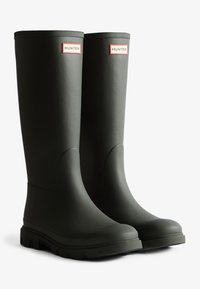 Bottes de pluie en caoutchouc vertes, montantes jusqu'aux genoux, avec une surface texturée et une semelle à crampons. Présente un logo "HUNTER" rouge et blanc à l'avant.