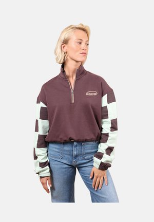 Femme portant un sweat-shirt violet à fermeture éclair trois-quarts avec des manches à carreaux et un jean bleu, regardant sur le côté sur un fond clair uni.
