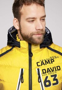 Żółta kurtka puchowa z granatowymi akcentami, posiada zip z przodu, dwie kieszenie na piersi oraz odważne czarne grafiki na materiale.