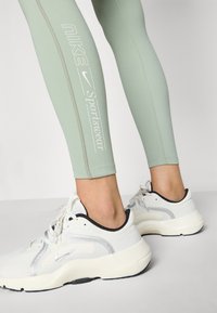 Világoszöld leggings fehér Nike logóval és "Sportswear" felirattal, fehér, fekete részletekkel és ezüst díszítéssel ellátott sportcipőkkel párosítva.