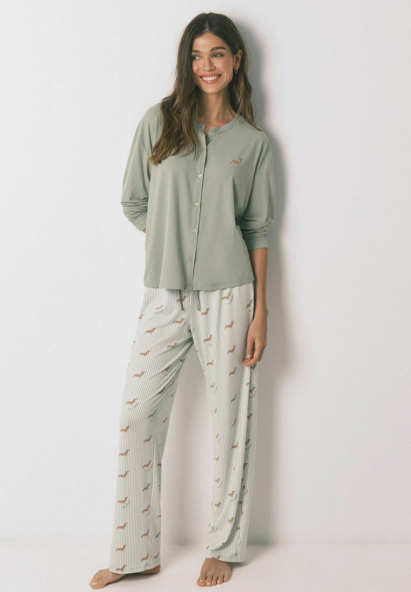 Woman smiling, wearing light green buttoned pajama top and white striped pajama pants with small brown dog patterns, standing barefoot.