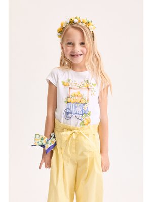Ragazza giovane che indossa una fascia per capelli a tema limone, una maglietta bianca con stampa di carretto di limoni, pantaloni gialli a vita alta e un fazzoletto da polso con motivo limone.