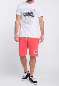 T-shirt bianca con stampa grafica nera e testo, abbinata a pantaloni corti corallo con un logo rotondo; il modello indossa sneakers bianche e nere.