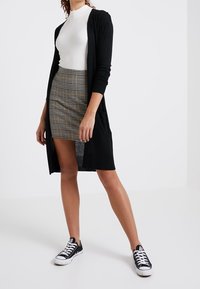 Svart lång cardigan över en vit ribbad topp, kombinerad med en rutig minikjol. Modellen bär svartvita sneakers.