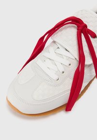 White sneaker with woven texture, red laces, and a gum outsole. Features rounded toe and padded tongue for comfort.
