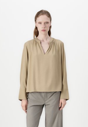 Blusa beige de manga larga con escote en V y detalles fruncidos en el cuello. La tela es suave y el corte es holgado y cuadrado.