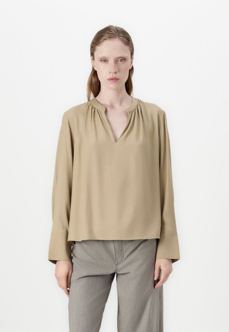 Blouse beige à manches longues avec un décolleté en V et des détails froncés au niveau du cou. Le tissu est lisse et la coupe est ample et carrée.