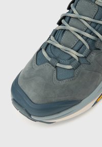 Γκρι αθλητικό παπούτσι με ανώτερη επιφάνεια από σουέτ και δίχτυ με υφή, κορδόνια σε ανοιχτό γκρι που κάνουν αντίθεση και στιβαρή λαστιχένια σόλα με πορτοκαλί διακριτικά σήματα.