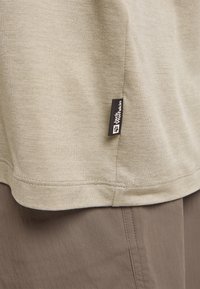 Beige Baumwoll-T-Shirt mit glatter Textur, ausgestattet mit einem genähten Saum und einem schwarzen Etikett mit weißem Text an der Seite.