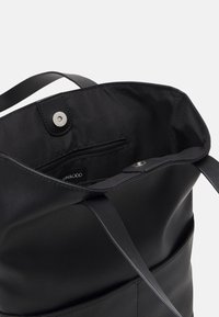 Sac cabas noir à surface extérieure lisse, ouverture sur le dessus et fermeture à pression. Intérieur avec une poche zippée et une doublure douce.