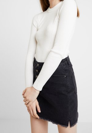 Femme portant un haut blanc côtelé à manches longues et une jupe en jean noire taille haute avec des boutons et un ourlet effiloché.