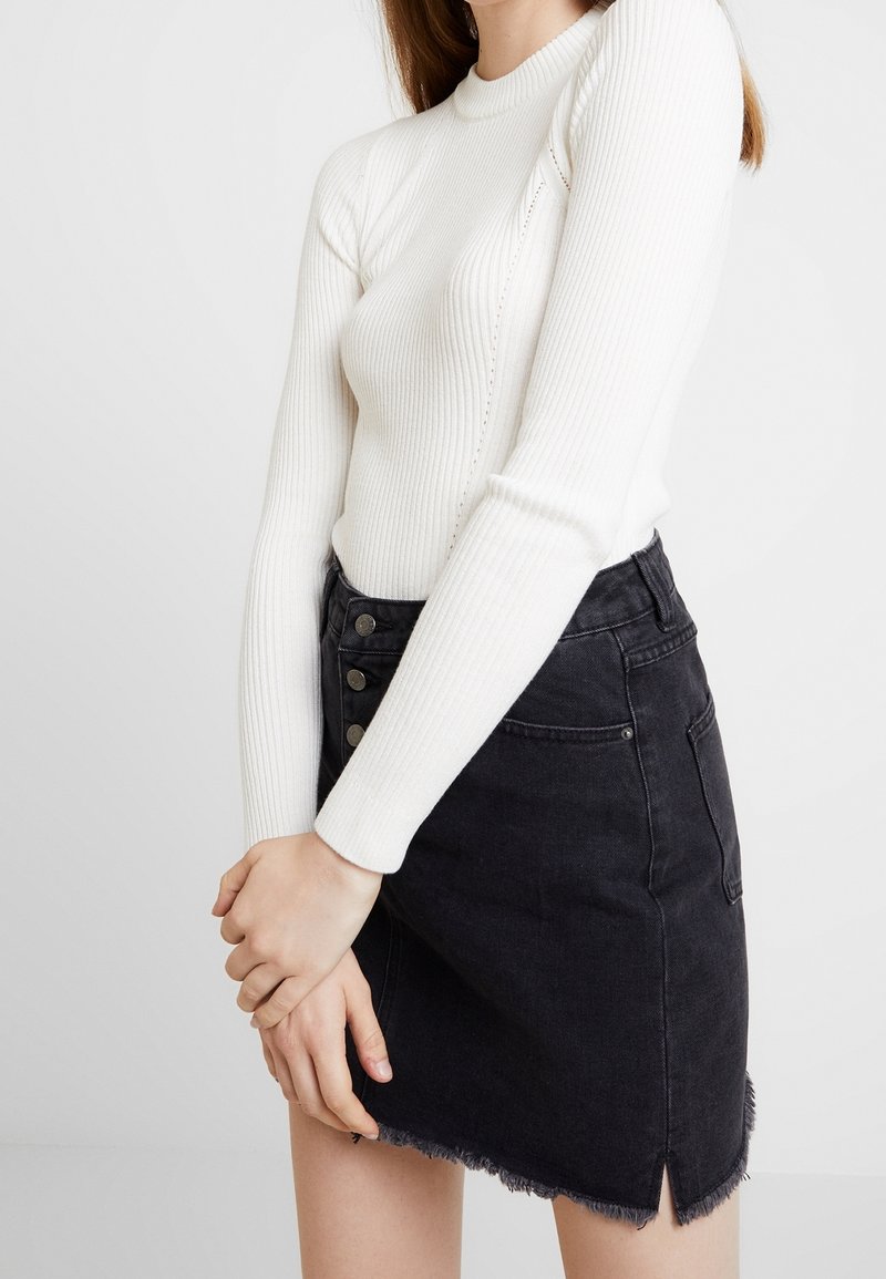 Femme portant un haut blanc côtelé à manches longues et une jupe en jean noire taille haute avec des boutons et un ourlet effiloché.