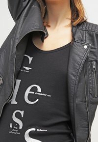 Osoba ubrana w czarną skórzaną kurtkę na czarną koszulę z srebrnym napisem "Założone w 1981, Seksowny, Młody, Przygodowy, Uwodzicielski."