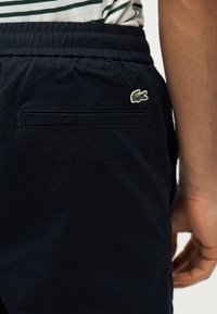 Κοντινό πλάνο ενός ατόμου που φοράει σκούρο μπλε παντελόνι με πράσινο λογότυπο κροκόδειλου κοντά στην πίσω τσέπη και ριγέ πουκάμισο μπουστάρει μέσα στο παντελόνι.