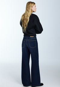 Vysoký pás, široké tmavé džínsy z denimu s kontrastným oranžovým prešitím a dvoma zadnými vreckami. Vrchný diel je čierna, voľne strihaná mikina.