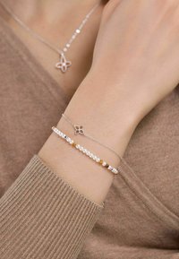 Bracciali d'argento su un polso: uno con un ciondolo floreale e accentuazioni di gemstone arancioni, l'altro con perline bianche e multicolori.