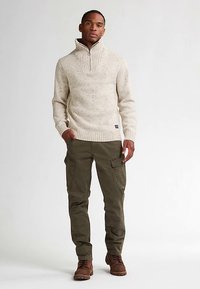 Beżowy sweter na drutach z wysokim kołnierzem, z fakturowanym wzorem. Noszony z oliwkowymi spodniami bojówkami i brązowymi butami. Prosty i praktyczny design.