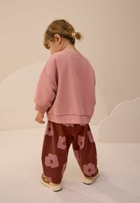 Ružový mikina s oversized strihom v kombinácii s hnedými voľnými nohavicami so vzorom ružových kvetov. Dieťa je otočené chrbtom, čo ukazuje účes.