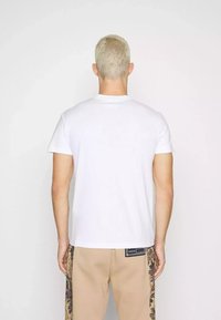 Maglietta di cotone bianca con collo rotondo, maniche corte e taglio dritto, mostrata di schiena. Indossata con pantaloni beige con un motivo laterale.