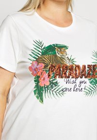Vit bomull t-shirt med en färgglad tiger-grafik omgiven av tropiska blad och blommor, med dekorativ text i paljetter.