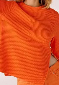 Pull tricot côtelé orange vif avec un détail de fermeture éclair sur le côté, assorti à un pantalon orange, porté par une personne aux cheveux blonds.