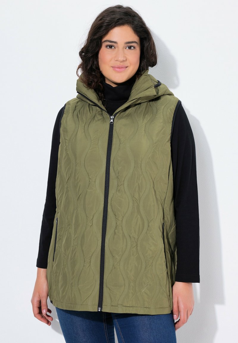 Gilet verde oliva trapuntato con cappuccio, dotato di zip frontale, tasche laterali e un motivo a onde testurizzato. Indossato sopra una maglietta nera a maniche lunghe.