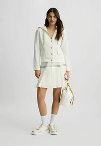 Weißer strukturierter Jacke mit Kapuze und goldenen Knöpfen, kombiniert mit einem weißen, gefältelten Rock mit schwarzem Saum und goldverzierten Sneakers.