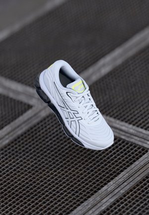 Scarpa sportiva bianca con tomaia testurizzata, suola in gomma nera e dettagli giallo neon sulla linguetta. Presenta il logo e i lacci.
