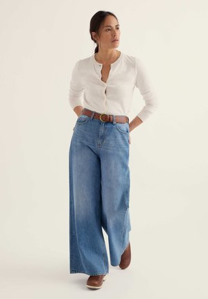 Naine seisab, kandes valget nööbitavat kardigani, laia jalaga siniseid teksaseid pruuni vöö ja pruunide kingadega, vaadates küljele.