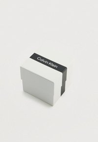 Weißer Karton mit einem schwarzen horizontalen Streifen, beschriftet mit "Calvin Klein." Der Karton hat eine glatte Textur und eine quadratische Form mit einem Deckel.