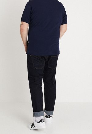 Granatowy t-shirt, ciemne dżinsy z podwiniętymi nogawkami i białe sneakersy z czarnymi akcentami. Prosty design o swobodnym kroju i widocznym przeszyciu.