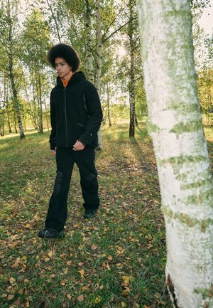 Chaqueta negra con cremallera y bolsillos, capa interior naranja, pantalones negros con parches texturizados en las rodillas, caminando sobre un terreno de hierba entre abedules.