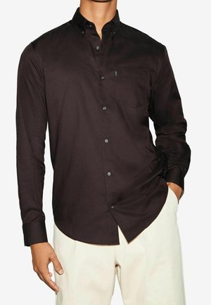 Homme portant une chemise marron foncé cintrée à manches longues avec une poche poitrine et un pantalon beige clair, debout avec une main dans la poche.
