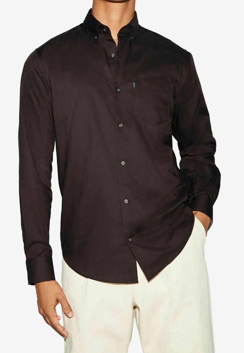 Homme portant une chemise marron foncé cintrée à manches longues avec une poche poitrine et un pantalon beige clair, debout avec une main dans la poche.