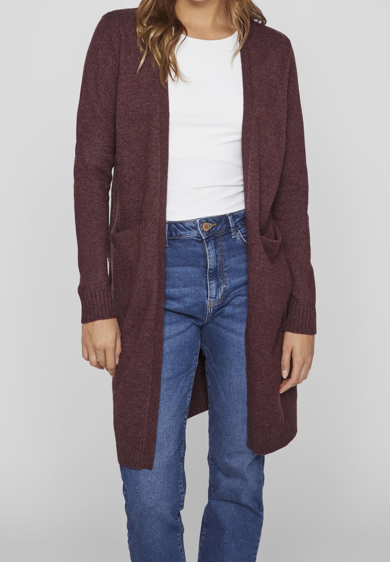 Femme portant un long cardigan grenat par-dessus un haut blanc et un jean bleu, debout devant un fond uni gris clair.