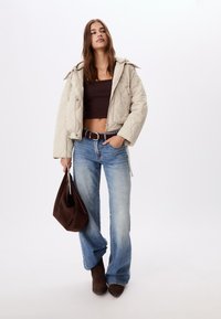 Veste matelassée beige avec fermeture éclair, crop top marron, jean flare bleu clair, ceinture marron, bottes en suède marron et sac marron.